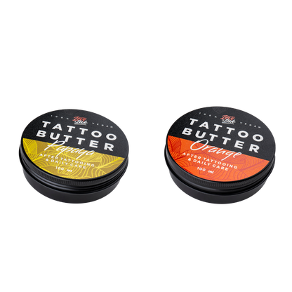 Two tins of Love Ink Tattoo Butter on a transparent background. One tin is labeled 'Orange' with an orange design, and the other is labeled 'Papaya' with an yellow design. Both tins are 100 ml and are intended for after tattooing and daily care.
