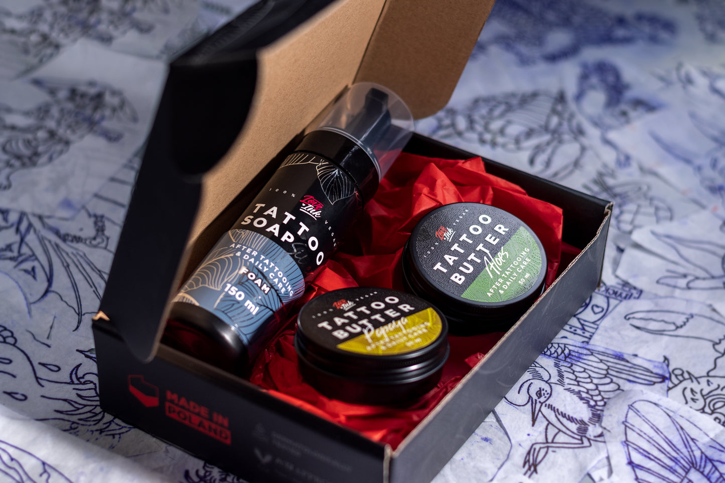 An open box containing Love Ink tattoo care products, including a bottle of Tattoo Soap and two tins of Tattoo Butter (Papaya and Aloe), placed on red tissue paper. The background features various tattoo sketches and designs
