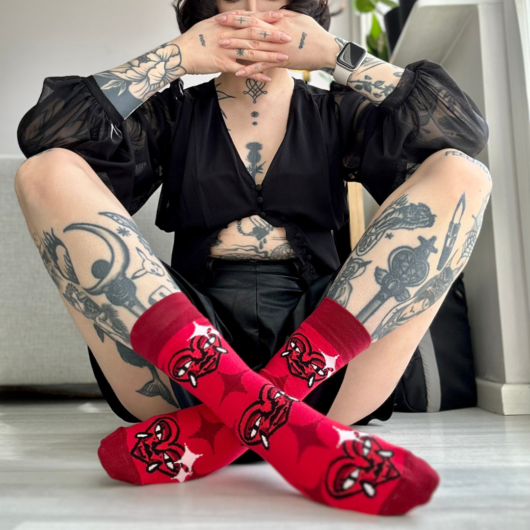 Tattooed person sitting on the floor with legs crossed, wearing red socks with a stylized heart design featuring horns and fangs, in a brightly lit indoor setting.