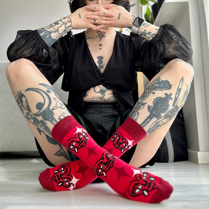 Tattooed person sitting on the floor with legs crossed, wearing red socks with a stylized heart design featuring horns and fangs, in a brightly lit indoor setting.