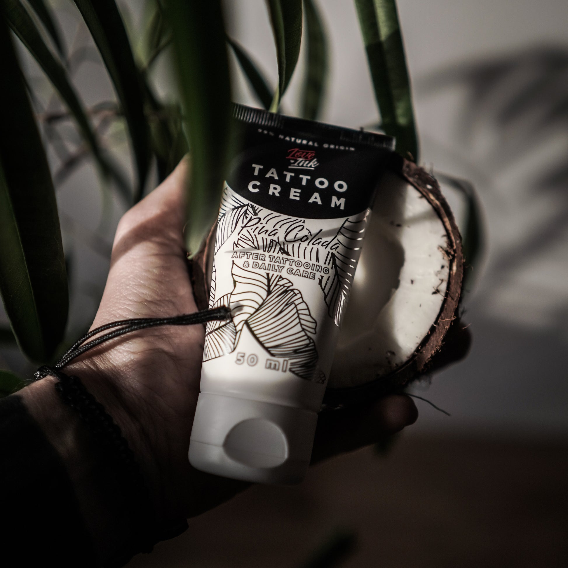 Hand holding a tube of Tattoo Cream by LoveInk with a split coconut in the background, emphasizing the product's natural ingredients.