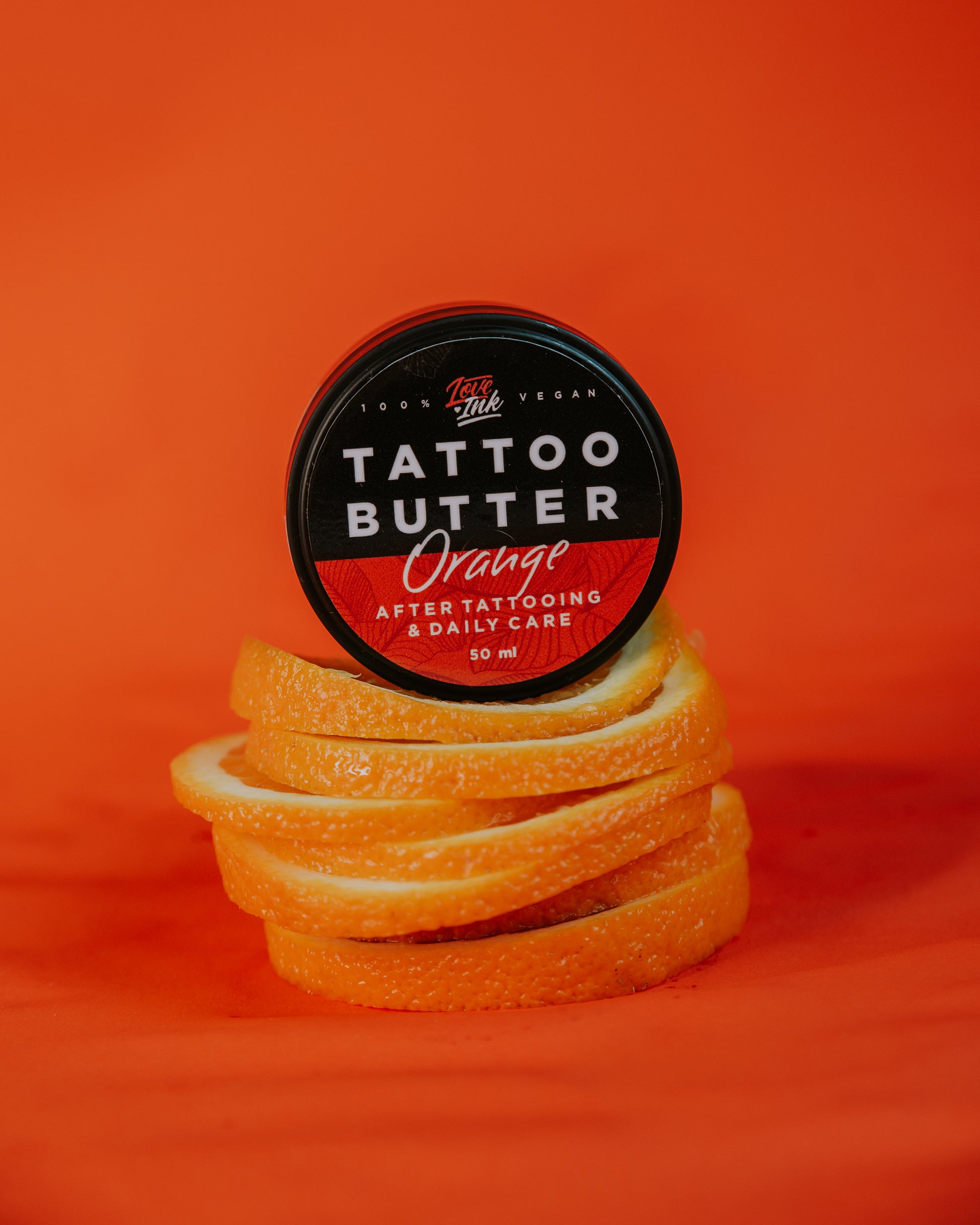 Jar of Tattoo Butter Orange by LoveInk surrounded by orange slices against an orange background, emphasizing the fresh, citrus scent of the product.