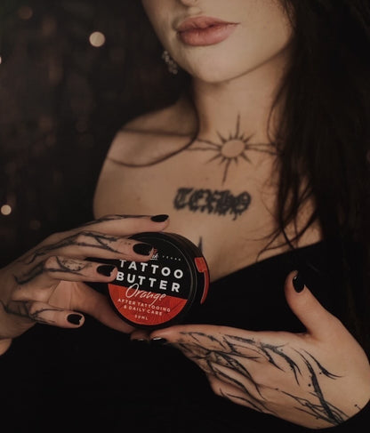 Close-up of a person with tattoos holding a jar of Tattoo Butter Orange by LoveInk, showcasing the product's relevance to tattoo care.