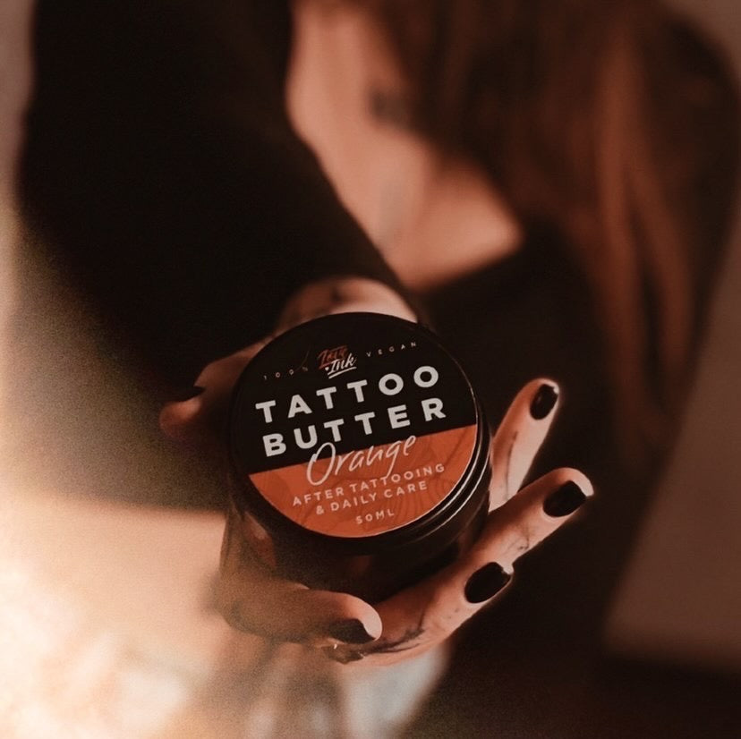 Person holding a jar of Tattoo Butter Orange by LoveInk in a dimly lit, warm-toned environment, highlighting the product's elegance and suitability for after tattooing care.