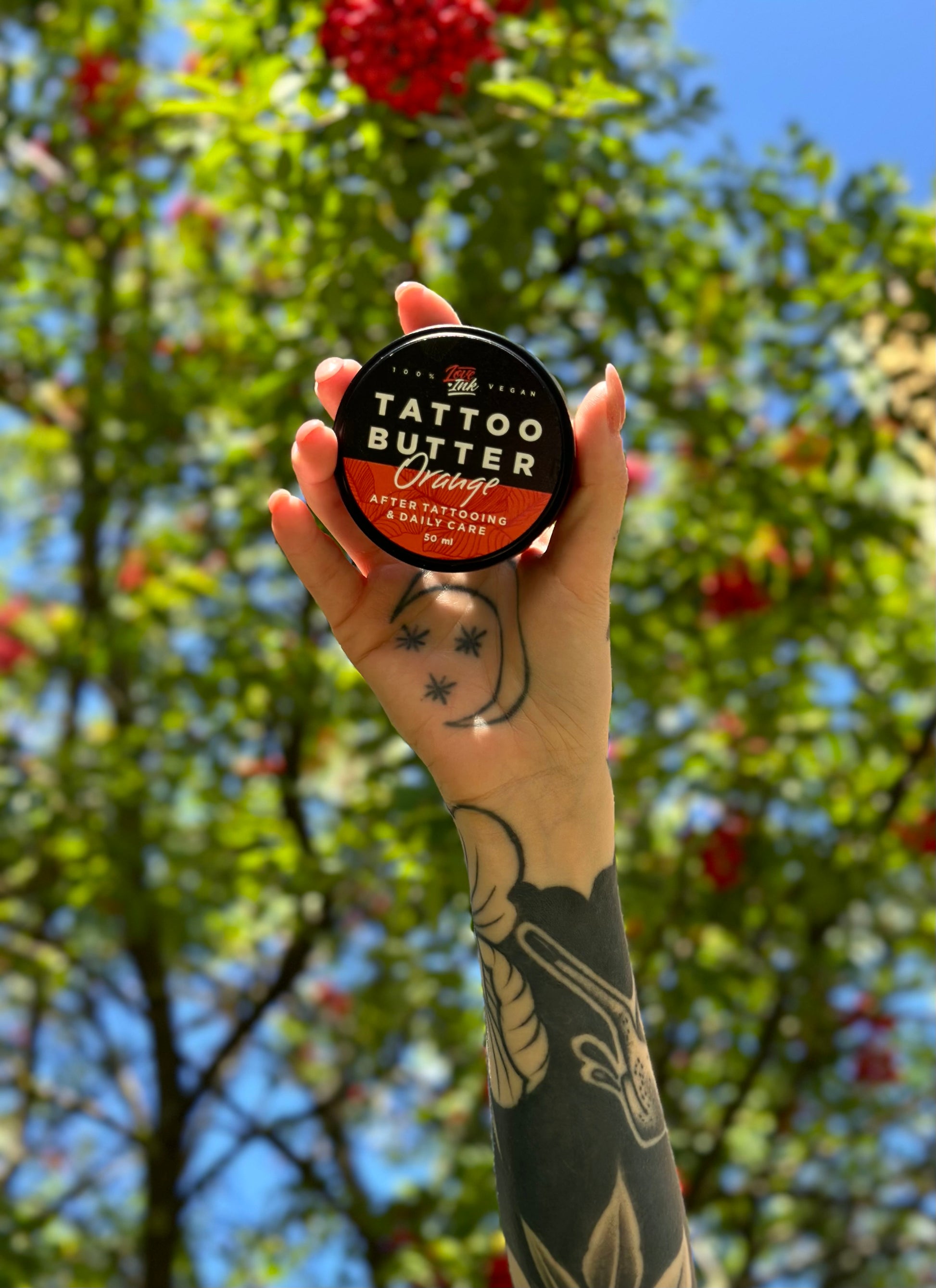 Hand holding a jar of Tattoo Butter Orange by LoveInk with a green tree and blue sky in the background. The image emphasizes the product's connection to nature.